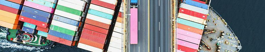 Description Aerial view of a container ship passing beneath a suspension bridge. Semi truck with pink cargo container crosses above.